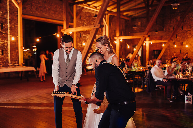ženích a nevesta s gitarou, nočná svadba v sklade, teplé zlaté osvetlenie, tanečný parket s hosťami, hudobný výkon na svadbe, romantická atmosféra s lampičkami. Karolinenhof Farm, Hainburg.