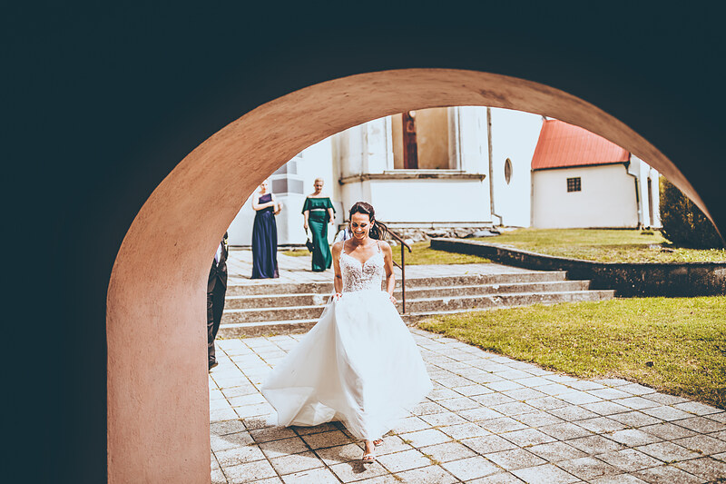nevesta v bielych šatách, terakotový oblúk, schodiská a trávnik, slnečný deň, svadobný sprievod, biela kaplnka v pozadí. Hacienda Šišov.