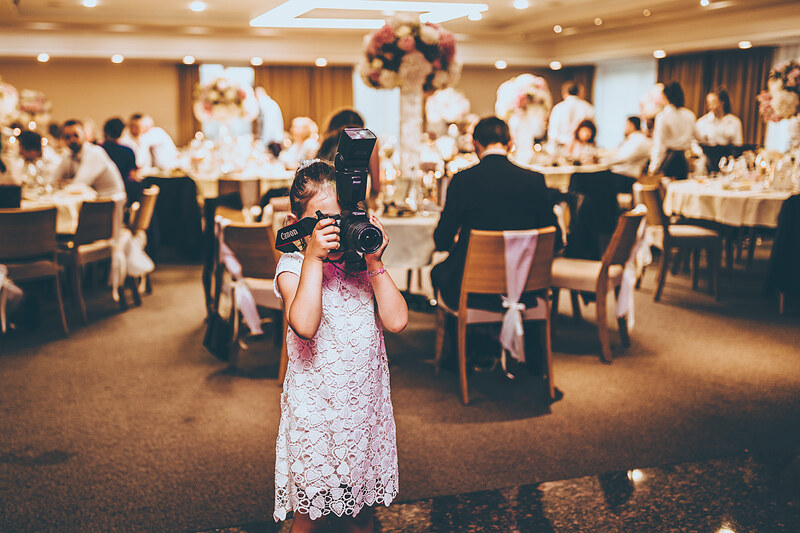 dievča s fotoaparátom, svadobná sála s hosťami, biele čipkované šaty, svadba v interiéri, stolový dekor s kvetmi, teplé osvetlenie v sále. Village Resort Hanuliak.