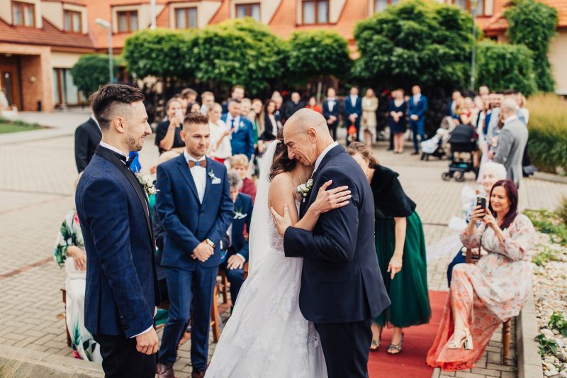 nevesta v bielych šatách, otec objíma nevestu, svadobný sprievod na nádvorí, hostia v modrých oblekoch, obrad na červenom koberci, slnečný deň vonku. Pivnica Radošina.