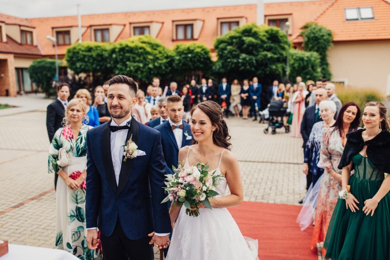 nevesta s kvetmi, ženích v obleku, pár drží ruky, hostia na dvore, červený behúň, tehlová budova v pozadí, denné svetlo, slávnostná atmosféra. Pivnica Radošina.