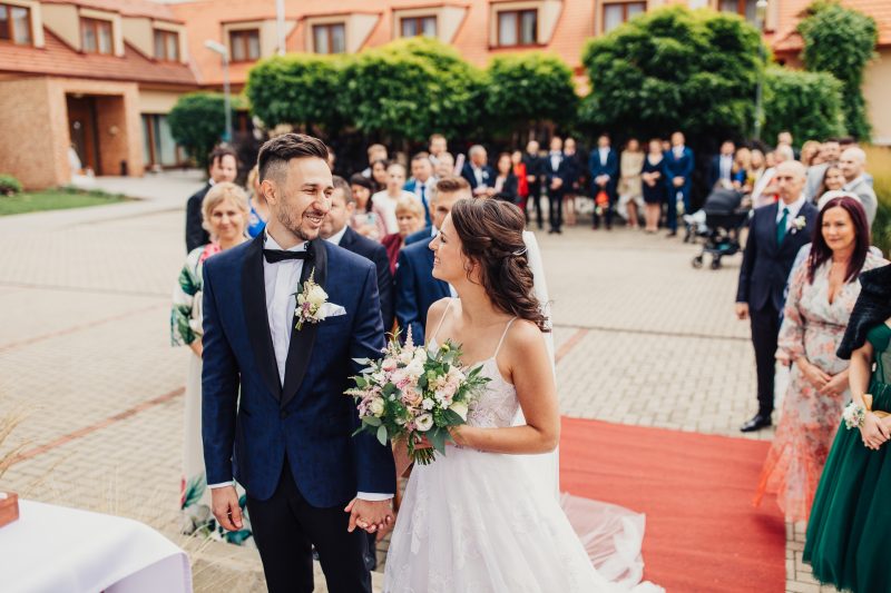 nevesta s kvetmi, ženích v modrom obleku, svadobný sprievod na dvore, červený behúň, hostia v pozadí, denné svetlo na nádvorí. Pivnica Radošina.