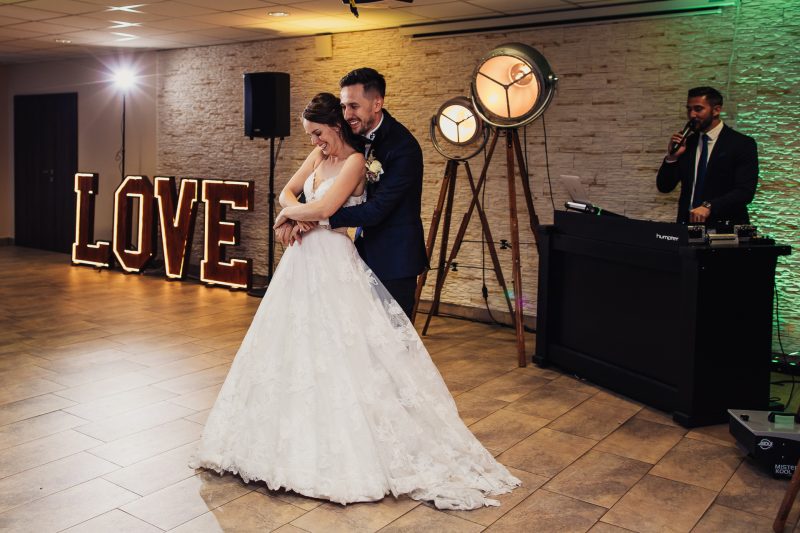 nevesta v bielych šatách, ženích v tmavom obleku, prvý tanec, neónový nápis love, industriálna sála s tehlovou stenou, teplé osvetlenie, pár tancuje spolu. Pivnica Radošina.