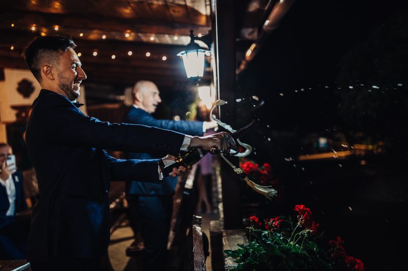 trubkár v noci, červené kvety pri lampe, večerná slávnosť vonku, modrá bunda a úsmev, nočné osvetlenie s dekoráciami, svadobný hosť hrá hudbu. Pivnica Radošina.