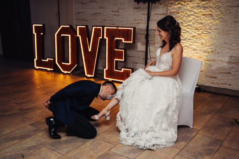 nevesta v bielych šatách, ženích kľačí pred nevestou, neónový nápis love, terakotová podlaha, teplé žlté svetlo, interiér s kamennou stenou. Pivnica Radošina.