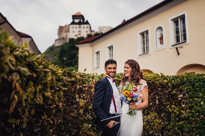 nevesta s kvetmi, ženích v obleku, vinič v pozadí, hrad na kopci, pár sa usmieva, krémová budova vzadu. Fatima, Trenčín.
