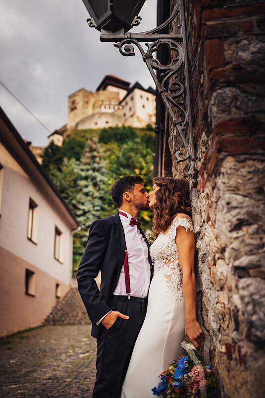 nevesta a ženích sa bozkávajú, pár v starej ulici, kamenná stena v pozadí, hrad na kopci, svadobný pár v kostýme, oblačné nebo nad mestom. Fatima, Trenčín.
