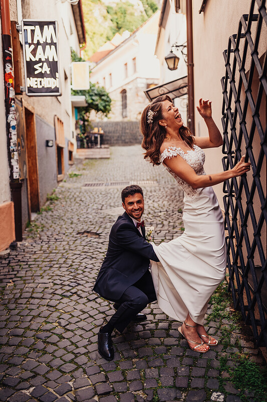 nevesta sa smeje v uličke, ženích drží nevestine šaty, pár v starej mestskej uličke, slnečné svetlo na dlažbe, radostná nevesta s kvetinami vo vlasoch, ženích v tmavom obleku drží nevestine šaty. Fatima, Trenčín.