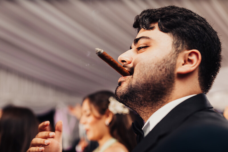 muž s cigarou v obleku, svadobný hosť fajčiaci, formálna svadobná udalosť, teplé svetlo na tvári, vážna nálada na svadbe, muž v čiernom obleku. Fatima, Trenčín.